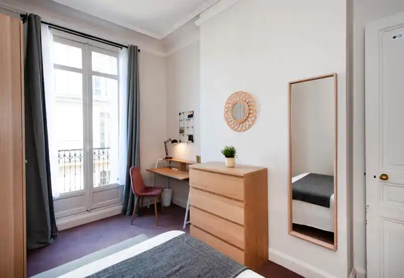 Bright, minimal bedroom with a large window, wooden dresser, full-length mirror and a cozy bed partially visible in the foreground. A small desk and chair by the window add a practical workspace.