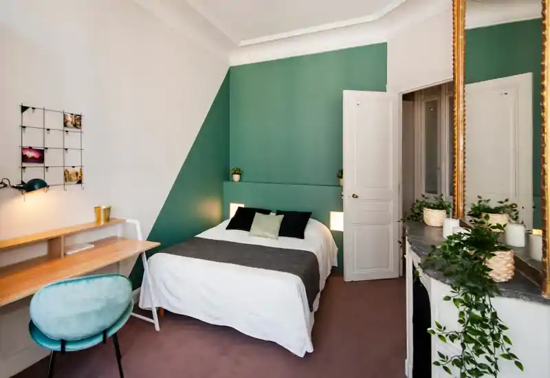 Chambre lumineuse et élégante avec un lit double en linge blanc, coussins décoratifs et chemin de lit sombre, un petit bureau en bois et un grand miroir orné sur une cheminée avec des plantes.