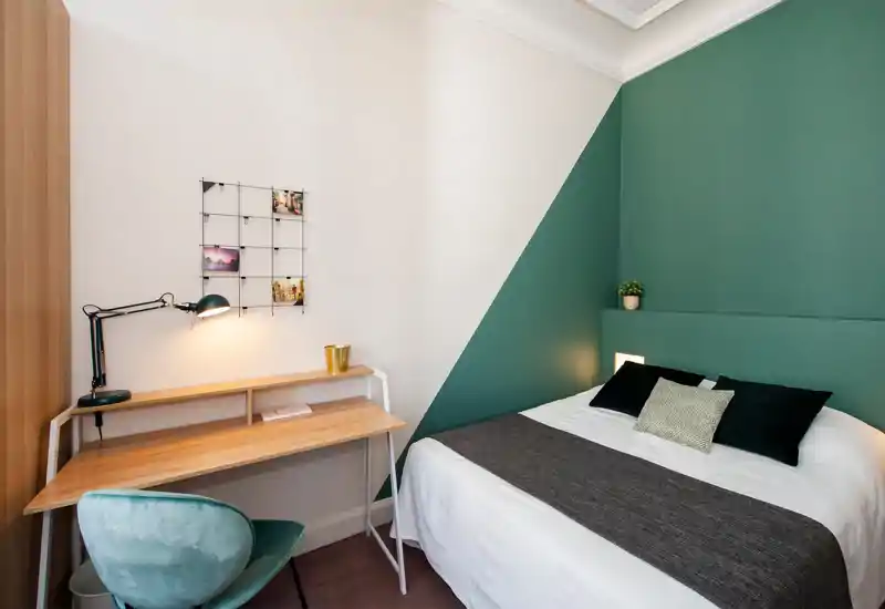 Chambre lumineuse et design avec un mur vert, lit double en linge blanc et coussins décoratifs, et un petit bureau en bois avec lampe de travail — idéale pour se détendre ou travailler.