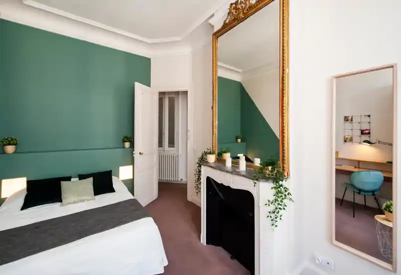 Chambre lumineuse et élégante avec un mur vert, lit double confortable, lampes de chevet et plantes décoratives ; grand miroir et coin bureau visibles.