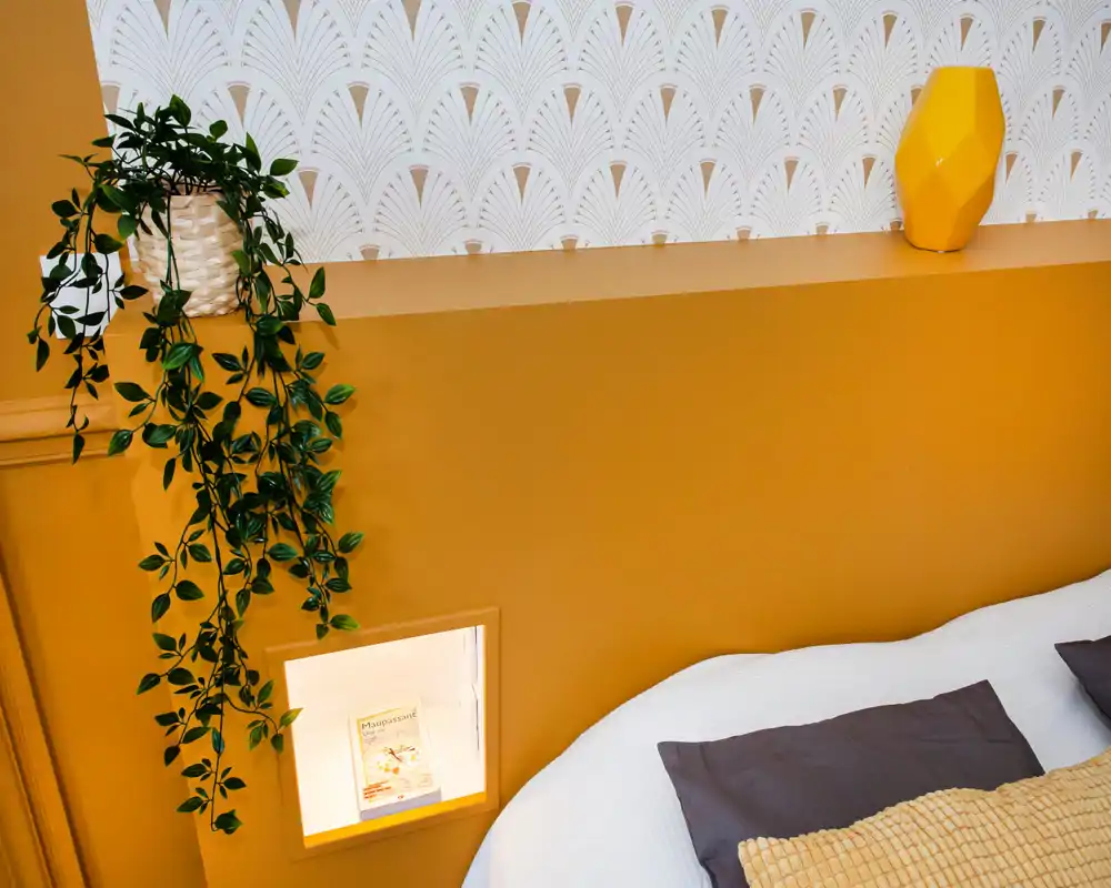 Coin de chambre lumineux et soigné avec tête de lit/étagère couleur moutarde, plante suspendue et lit bien fait avec coussins texturés — moderne, chaleureux et bien composé.