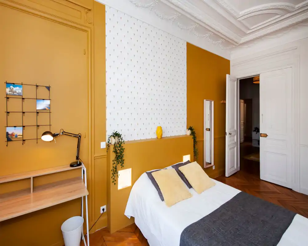 Chambre lumineuse et élégante avec un lit double en linge blanc, coussins moutarde et une niche décorative en tête de lit ; plafond orné et parquet chaleureux pour une atmosphère cosy et moderne.