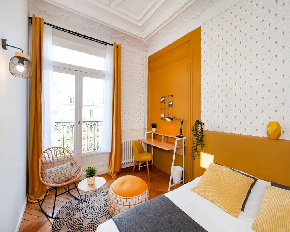 Chambre lumineuse et élégante aux accents moutarde : lit bien fait avec coussins texturés, petit bureau et coin détente près d'une grande fenêtre qui inonde la pièce de lumière naturelle.