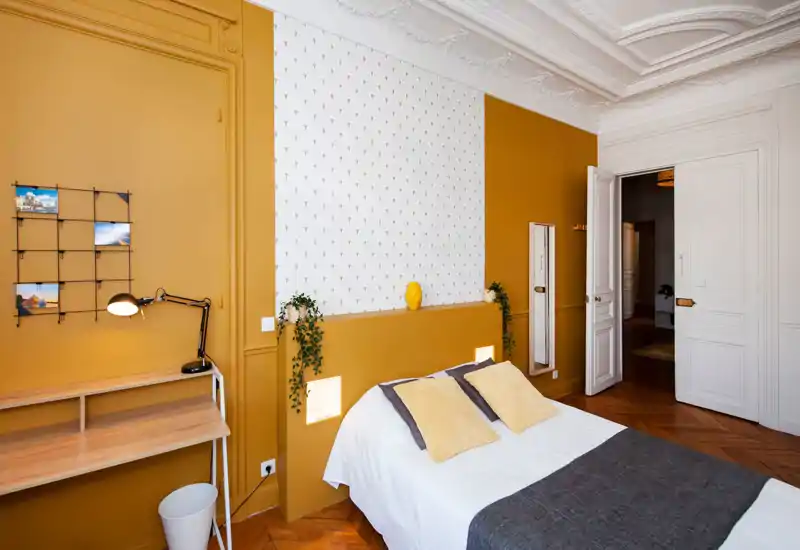 Chambre lumineuse et élégante avec un lit double en linge blanc, coussins moutarde et une niche décorative en tête de lit ; plafond orné et parquet chaleureux pour une atmosphère cosy et moderne.