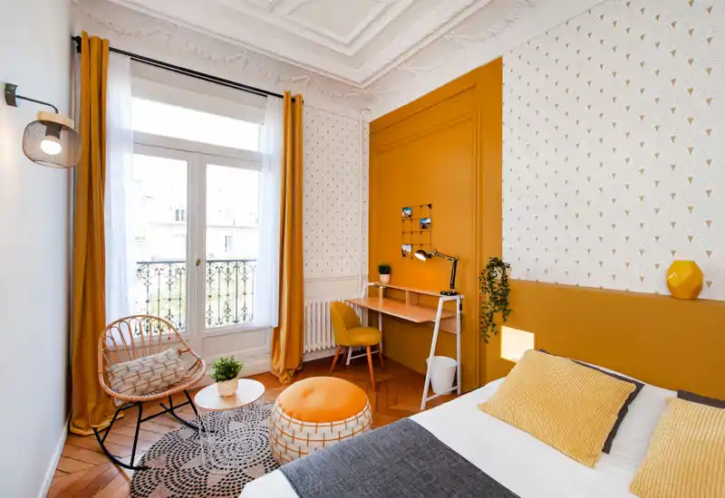 Chambre lumineuse et élégante aux accents moutarde : lit bien fait avec coussins texturés, petit bureau et coin détente près d'une grande fenêtre qui inonde la pièce de lumière naturelle.