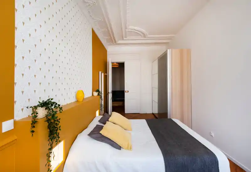 Chambre lumineuse et stylée avec un grand lit en linge blanc, coussins jaunes texturés et un couvre-lit foncé. La pièce offre des moulures décoratives, un mur d'accent à motif et une niche avec plantes pour une atmosphère chaleureuse et moderne.