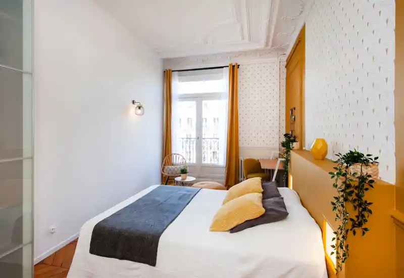Chambre lumineuse et élégante avec un grand lit en linge blanc, accents jaune moutarde et coussins décoratifs. La pièce dispose d'une grande fenêtre avec voilages, un coin salon, des plantes et un mur à motif pour une ambiance chaleureuse et moderne.