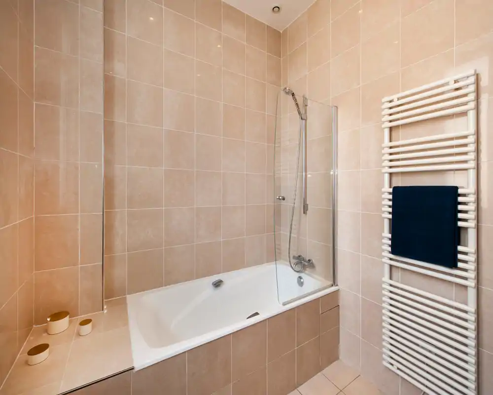 Salle de bain moderne carrelée avec baignoire intégrée, douchette et pare-douche en verre; sèche-serviettes avec une serviette foncée pour une touche pratique.