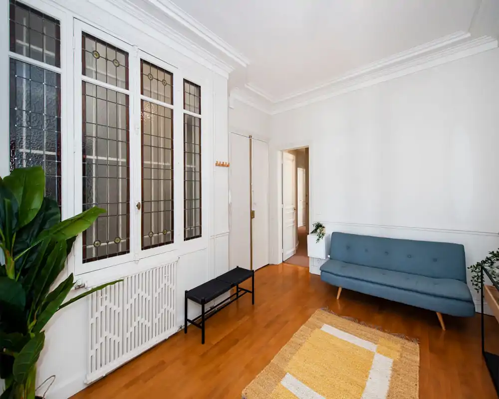 Pièce à vivre lumineuse et bien aménagée avec un canapé style mid-century, tapis décoratif et plantes. Grandes fenêtres à vitraux et moulures blanches apportent du caractère et une sensation d'espace.