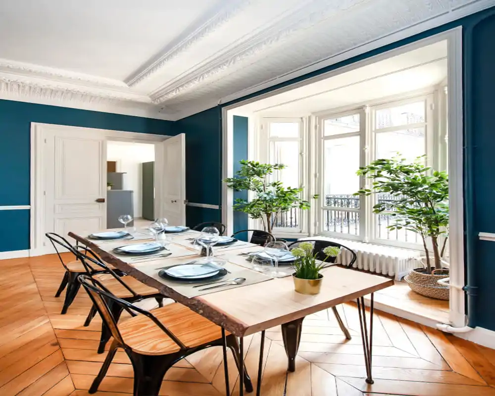 Salle à manger lumineuse et élégante avec une table en bois à chant vif pour six personnes, grandes fenêtres et plantes apportant une ambiance aérée.
