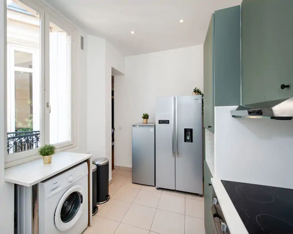 Cuisine moderne et lumineuse avec réfrigérateur en acier inoxydable, lave-linge, plaque à induction et placards verts ; agencement soigné et lumière naturelle par une grande fenêtre.