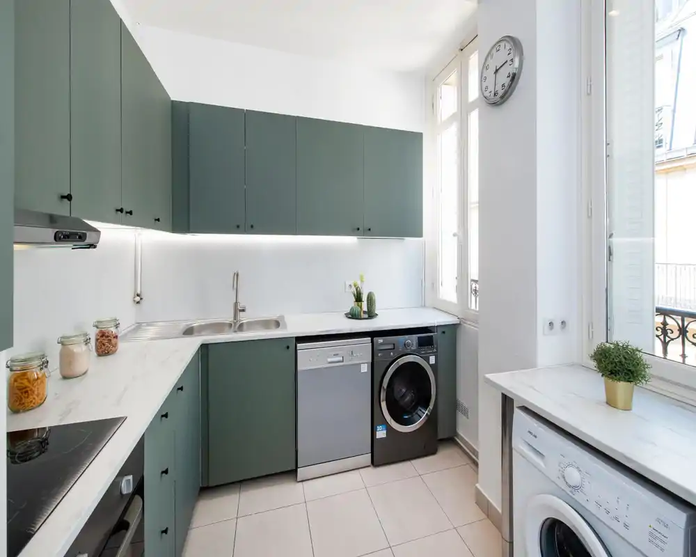 Cuisine moderne et lumineuse avec des placards verts, plans de travail effet marbre, électroménagers encastrés et grandes fenêtres offrant une belle lumière naturelle.