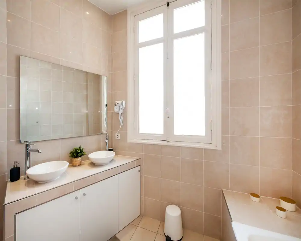 Salle de bains moderne et lumineuse avec vasques doubles, grand miroir et carrelage beige neutre ; espace propre et bien éclairé, idéal pour mettre en valeur les équipements contemporains.