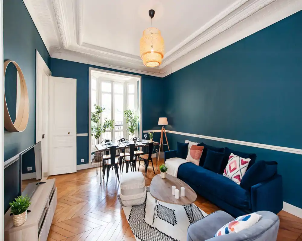 Salon lumineux et élégant avec canapé bleu profond, coussins décoratifs, tapis à motifs et table basse en bois ; large baie vitrée et coin repas visibles, ambiance chic d'appartement citadin.
