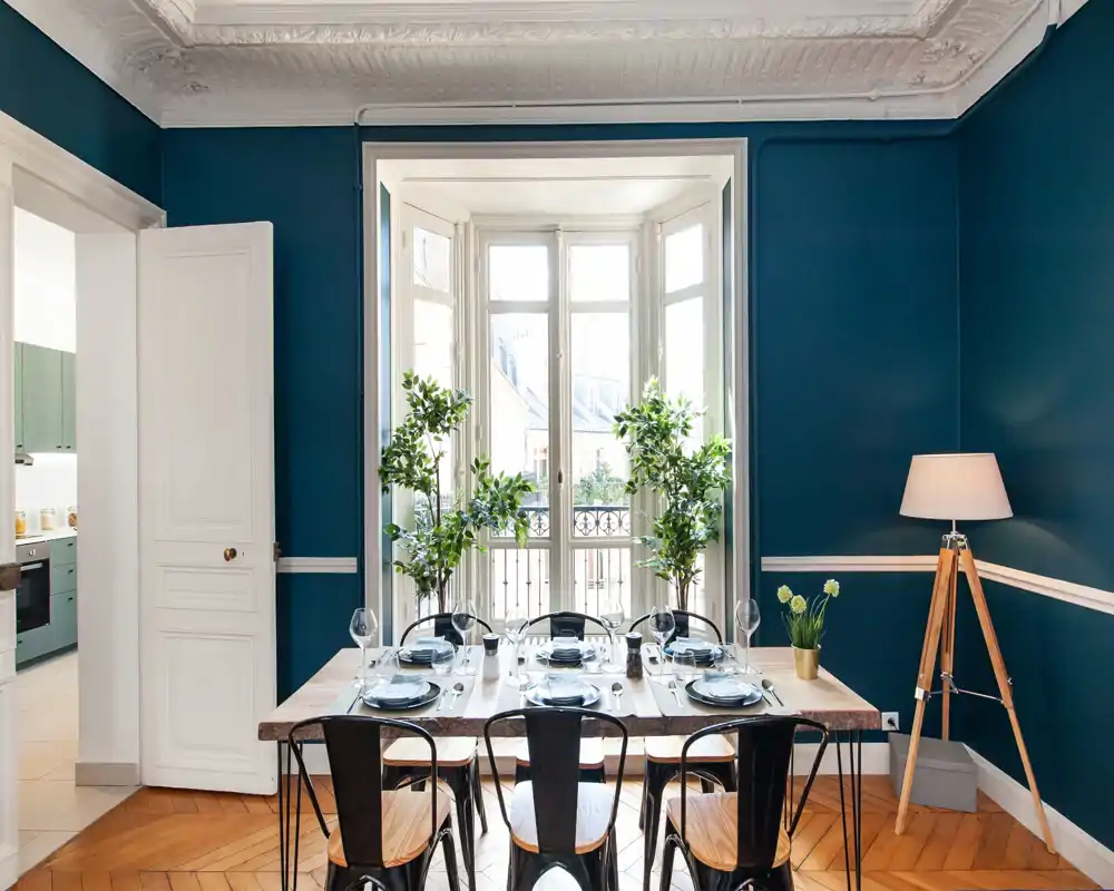 Salle à manger élégante avec une grande table en bois naturel dressée pour six, murs bleu canard, lumière naturelle abondante depuis une baie vitrée, plantes décoratives et lampe trépied pour une ambiance chaleureuse et stylée.