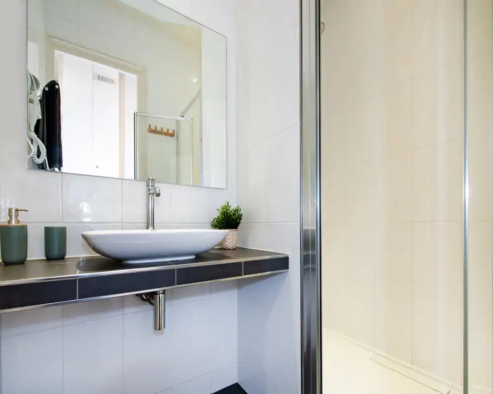 Salle de bains moderne et lumineuse avec vasque, grand miroir et porte de douche en verre ; carrelage blanc propre et petite tablette avec accessoires verts.