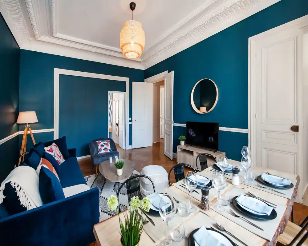 Salon élégant aux murs bleu profond, canapé confortable et table à manger dressée pour quatre — lumineux, bien mis en scène et parfait pour recevoir.