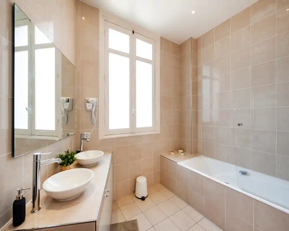 Salle de bains moderne et lumineuse avec baignoire, pare-douche en verre, doubles vasques et grand miroir ; carrelage beige neutre et grande fenêtre apportent une belle lumière naturelle.