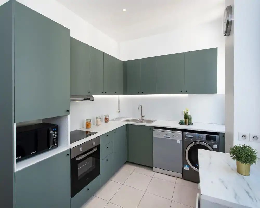 Cuisine moderne en L avec placards verts mats, four et lave-vaisselle intégrés, évier double et plans de travail effet marbre. Lumineuse et soignée, avec bocaux décoratifs et petites plantes apportant de la chaleur.