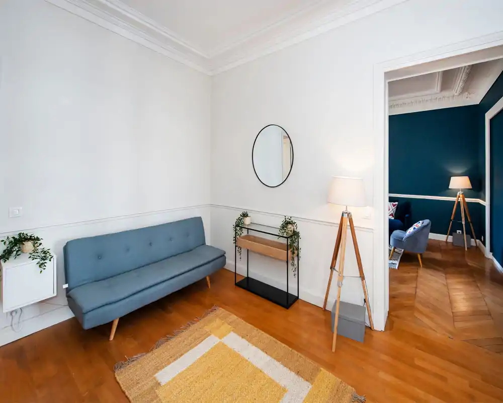 Espace de vie lumineux et moderne avec un canapé bleu style mid-century, parquet chaleureux et tapis décoratif ; petite console, plantes et lampe trépied donnent une ambiance accueillante.