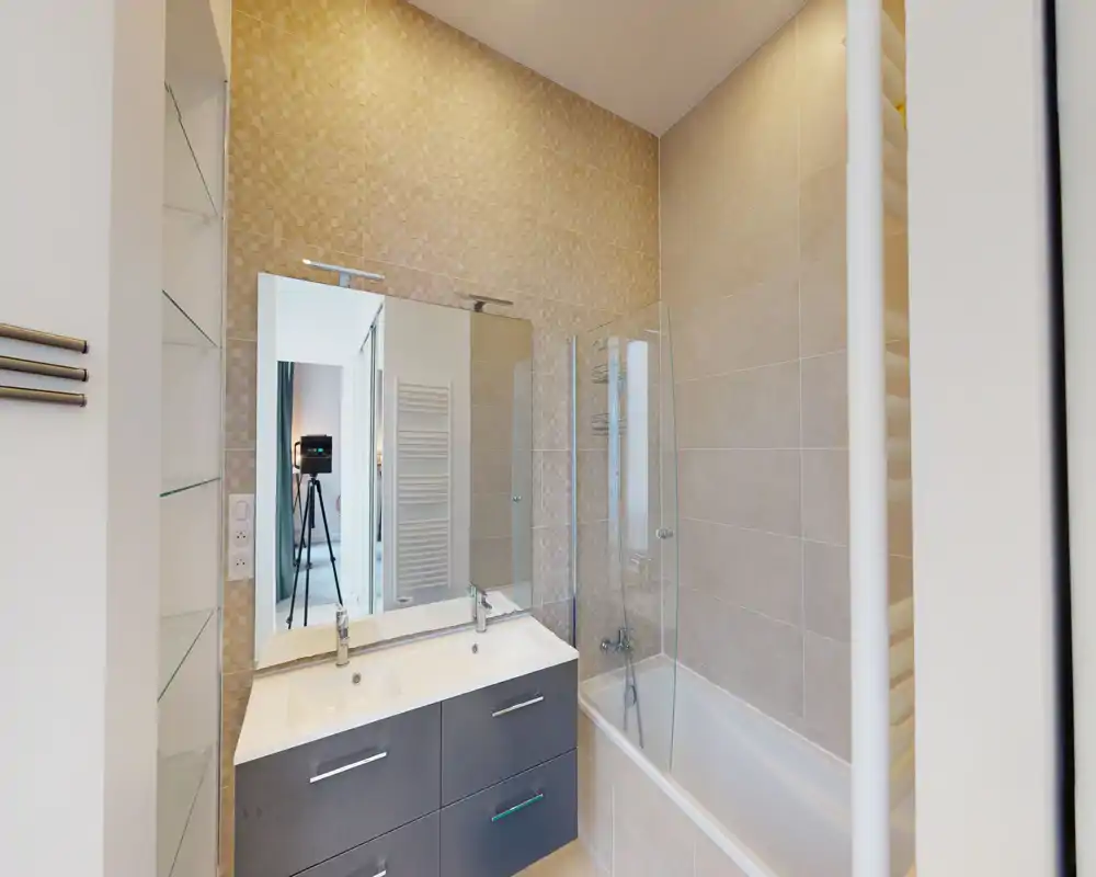 Salle de bains moderne avec vasque double, grand miroir et baignoire-douche avec pare-douche en verre ; carrelage beige neutre et équipements contemporains.