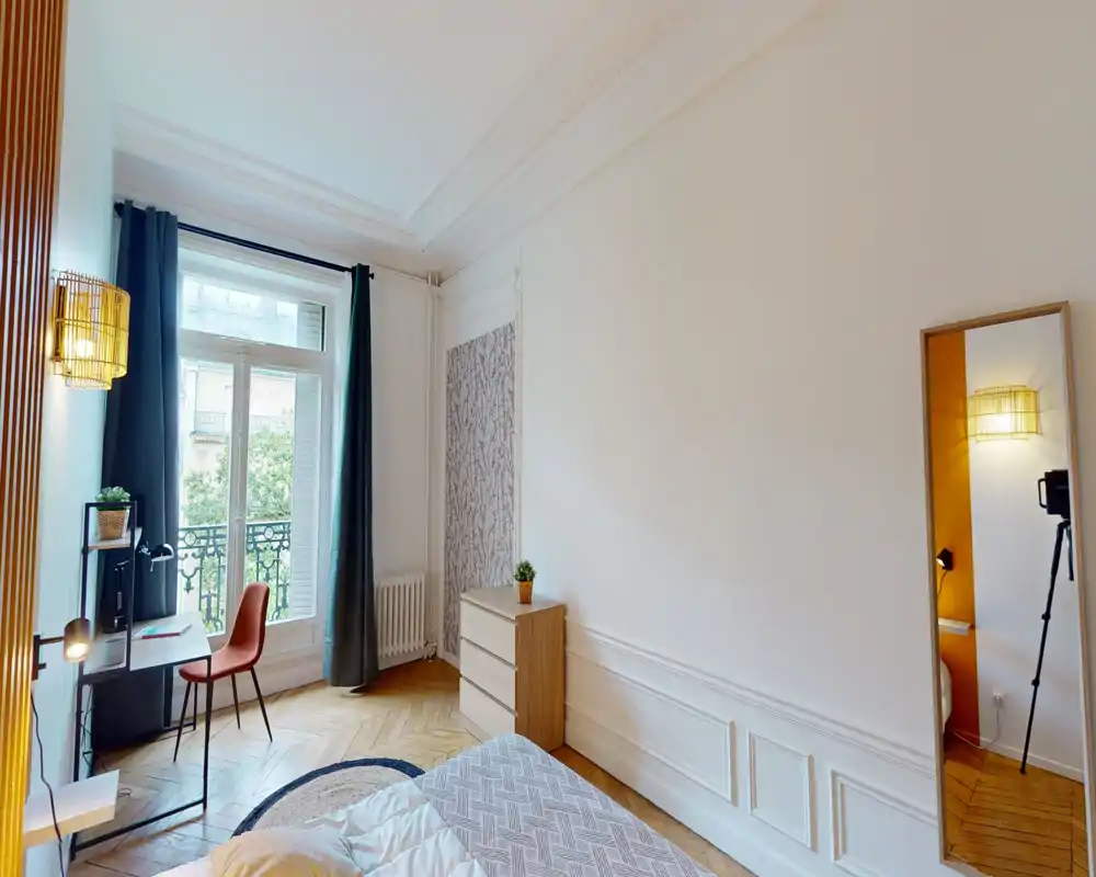 Chambre lumineuse et élégante avec hauts plafonds, parquet et lit confortable au premier plan. Bureau près de la fenêtre, lampe décorative et miroir en pied offrent un espace à la fois stylé et fonctionnel, parfait pour la location saisonnière.