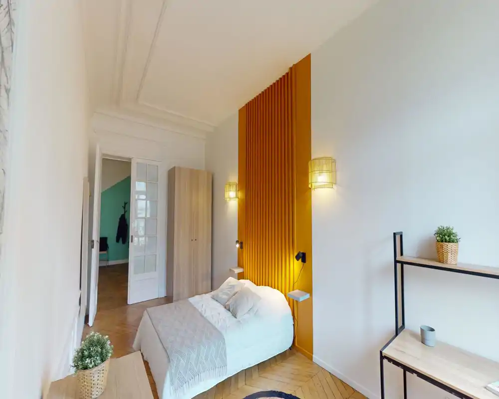 Chambre lumineuse et stylée avec lit double devant un panneau vertical couleur moutarde, appliques chaleureuses et parquet en bois — cosy et bien éclairée.