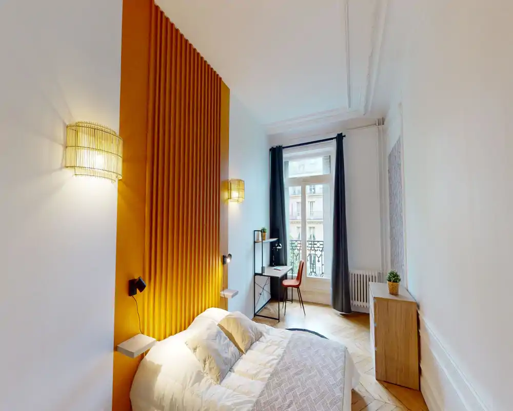 Chambre lumineuse et stylée avec un mur d'accent moutarde, lit double confortable et lampes de chevet ; bureau près de la fenêtre pour travailler à la lumière du jour.