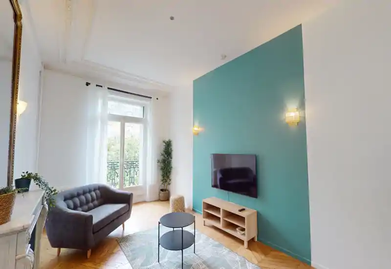 Salon lumineux et élégant avec mur d'accent vert-bleu, télévision murale, petit canapé et lumière naturelle provenant d'une grande fenêtre ouvrant sur un balcon.