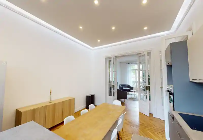 Salle à manger moderne et lumineuse avec une grande table en bois, chaises blanches et parquet, s'ouvrant sur un coin salon via des portes vitrées.