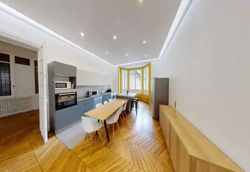 Cuisine moderne et spacieuse avec électroménagers intégrés, une grande table et une baie vitrée lumineuse. Le parquet en chevron et les meubles aux tons neutres offrent un espace élégant et chaleureux, parfait pour cuisiner et recevoir.