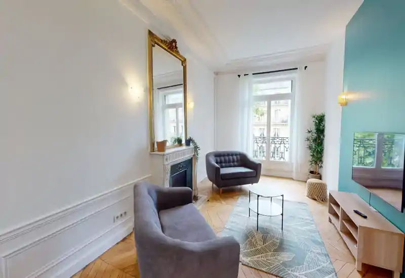 Bright, elegant living room with two comfortable grey armchairs, decorative rug, wall-mounted TV and a large window overlooking a balcony—ideal for relaxing.