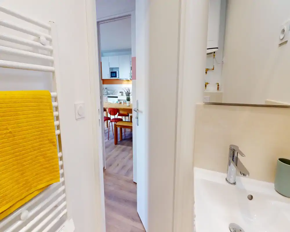 Bright, modern bathroom view with a white sink and chrome faucet in the foreground, a heated towel rail with a yellow towel on the left, and a doorway opening to the kitchen/dining area.