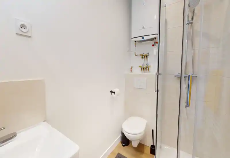 Compact modern bathroom with a wall-mounted toilet, corner shower with glass door and a small sink; clean white tiles and warm wood-look flooring create a tidy, functional space.