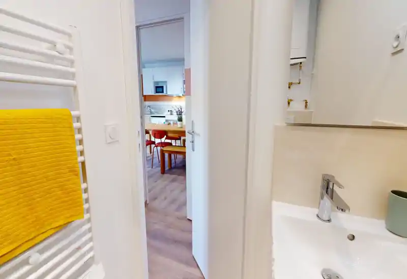 Bright, modern bathroom view with a white sink and chrome faucet in the foreground, a heated towel rail with a yellow towel on the left, and a doorway opening to the kitchen/dining area.