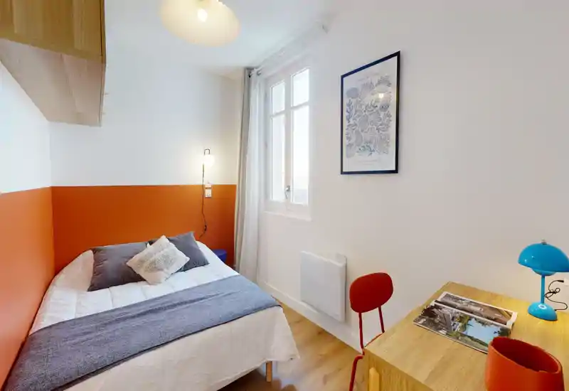 Compact, well-styled bedroom with a double bed against a warm orange accent wall, bedside lamp and a small desk by a bright window — ideal for a cozy city stay.