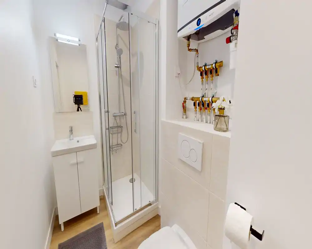 Salle de bains moderne et compacte avec cabine de douche d'angle en verre, plaque de chasse murale et petit meuble-lavabo ; murs blancs et sol effet bois pour un rendu propre et minimaliste.