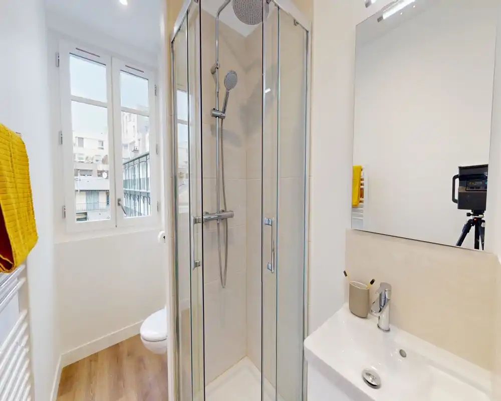 Salle de bain moderne et lumineuse avec cabine de douche d'angle en verre, miroir mural et petit lavabo — la lumière naturelle de la fenêtre et le sol effet bois apportent une atmosphère fraîche et accueillante.