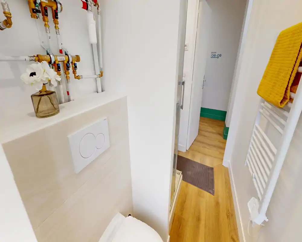 Coin salle de bains compact et moderne avec plaque de chasse d'eau, radiateur-serviette et porte vers le couloir ; carrelage blanc et sol effet bois pour un espace propre et fonctionnel.