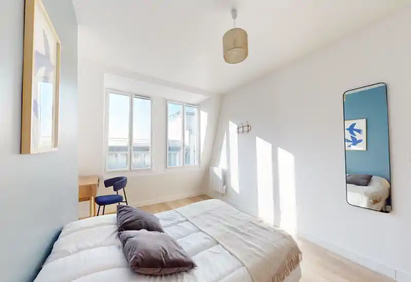 Chambre moderne et lumineuse avec un lit double bien fait, oreillers moelleux et un léger plaid. Grandes fenêtres apportant beaucoup de lumière naturelle et un petit bureau avec chaise près de la fenêtre.
