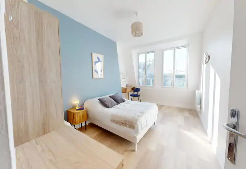 Chambre moderne et lumineuse avec un grand lit près d’un mur bleu, table de chevet et petit bureau près de la fenêtre — idéale pour un séjour reposant.