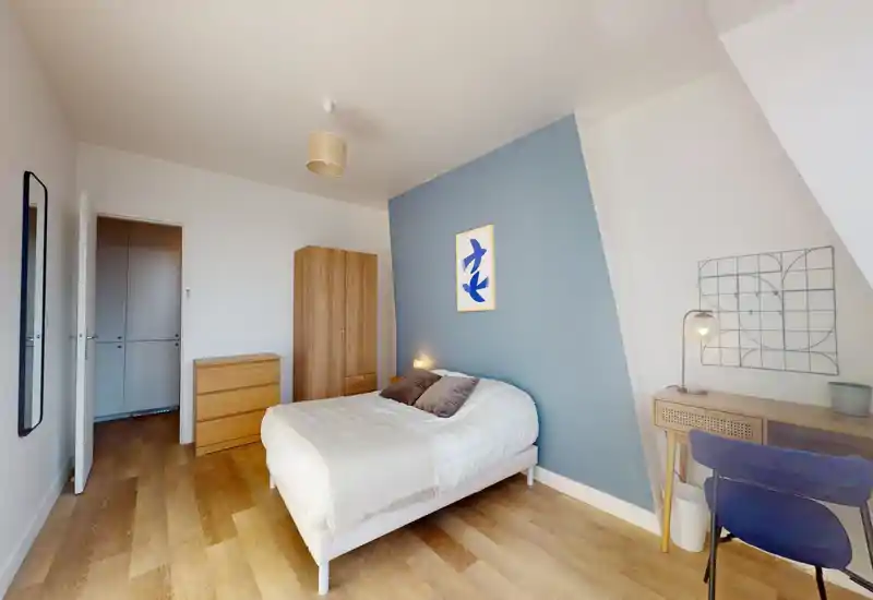 Chambre lumineuse bien mise en scène avec lit double devant un mur bleu clair, parquet et un petit espace de travail avec lampe — confortable et moderne.