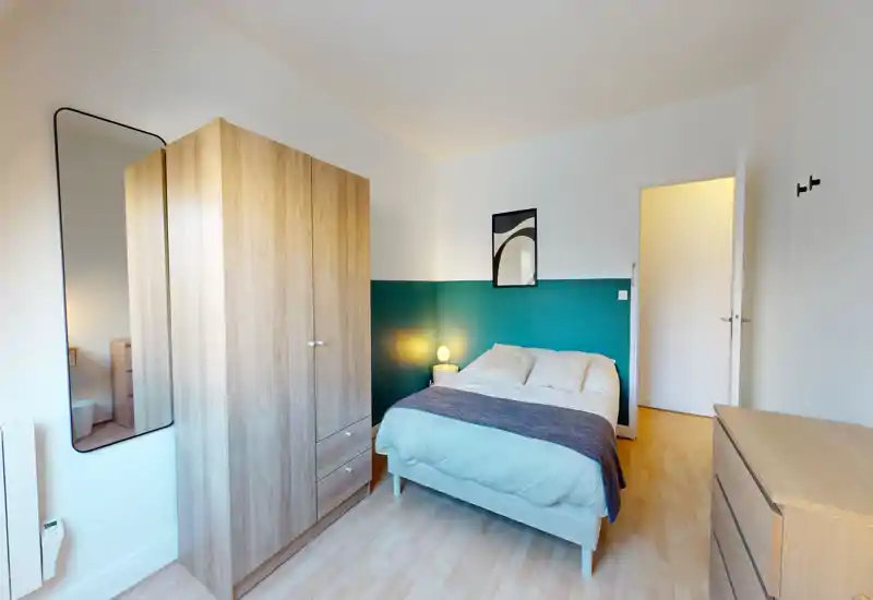 Chambre lumineuse et bien mise en scène avec un lit double devant un mur accent turquoise, lampe de chevet, armoire et commode — parfait pour une annonce locative.