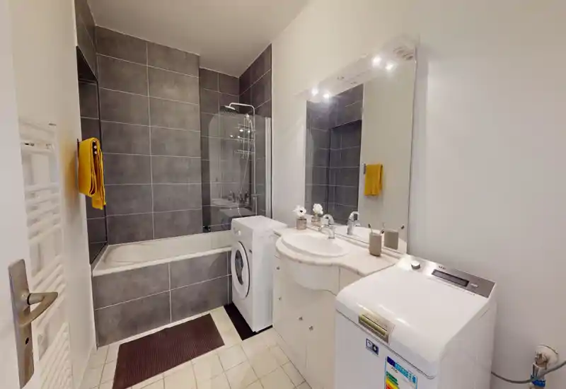 Salle de bains moderne et lumineuse avec baignoire/douche, murs carrelés, lavabo avec meuble et une machine à laver bien placée pour le linge.