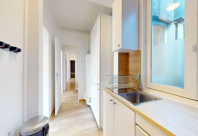 Kitchenette compacte et moderne avec placards blancs, plan de travail en bois et évier près d'une fenêtre lumineuse — agencement efficace pour un studio.