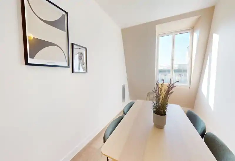 Coin repas lumineux et compact avec une table en bois clair, chaises rembourrées et une plante décorative au centre. La lumière naturelle par la fenêtre et les cadres muraux simples complètent cet espace moderne et accueillant.