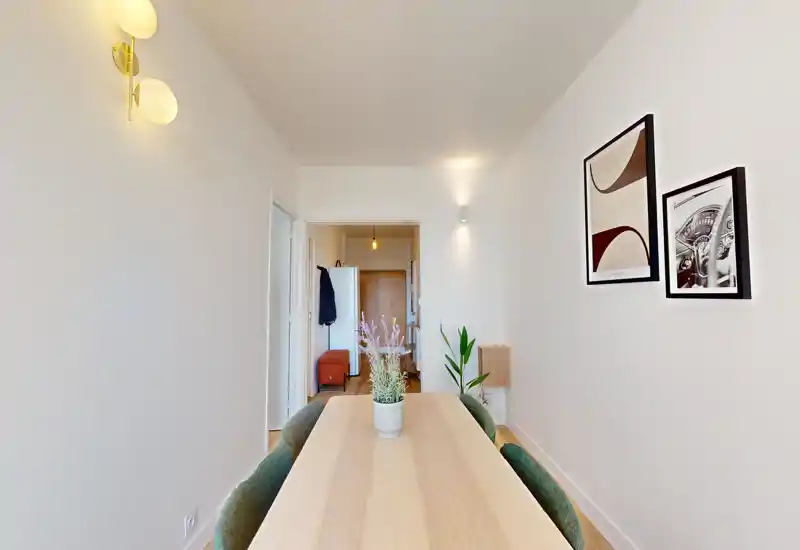 Coin repas moderne et lumineux avec table en bois clair, chaises vertes et cadres muraux; décoré d’un petit centre de table et d’un éclairage mural chaleureux.