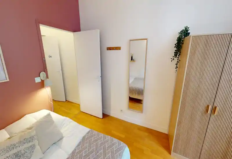 Camera da letto luminosa e accogliente con letto ben rifatto, plaid e cuscini decorativi, specchio a tutta altezza e armadio alto — pavimento in legno chiaro e parete rosa creano un’atmosfera calda.