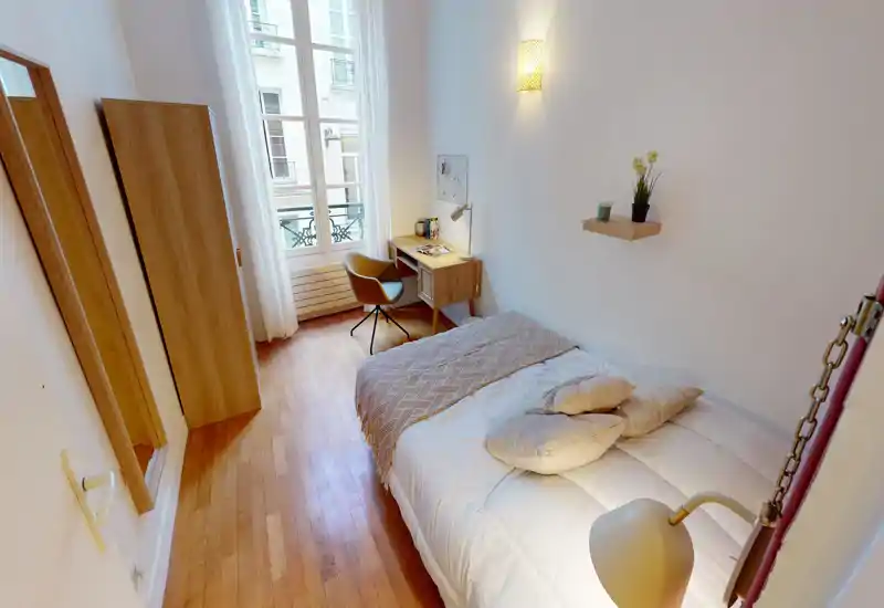 Camera da letto singola luminosa e ben arredata con letto comodo, pavimento in legno e scrivania vicino a una finestra alta che lascia entrare molta luce naturale.