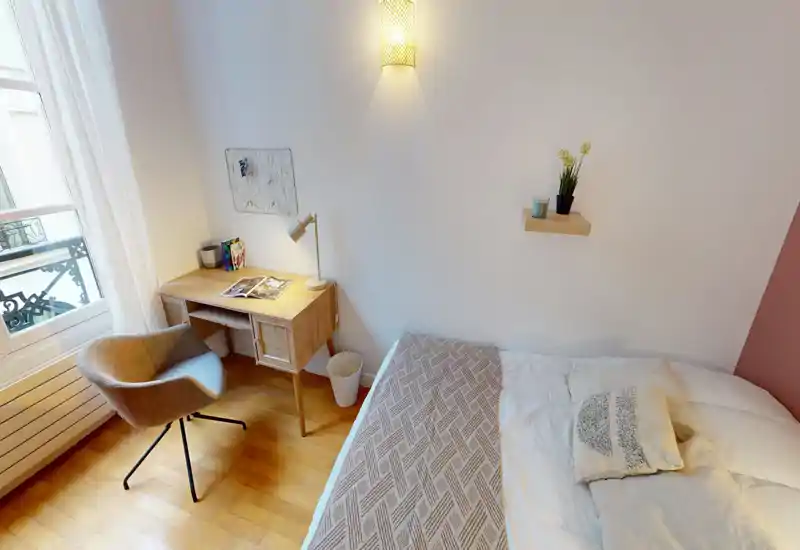 Camera da letto luminosa e accogliente con un letto comodo, coperta decorativa e una piccola scrivania in legno vicino alla finestra — perfetta per lavoro o relax.